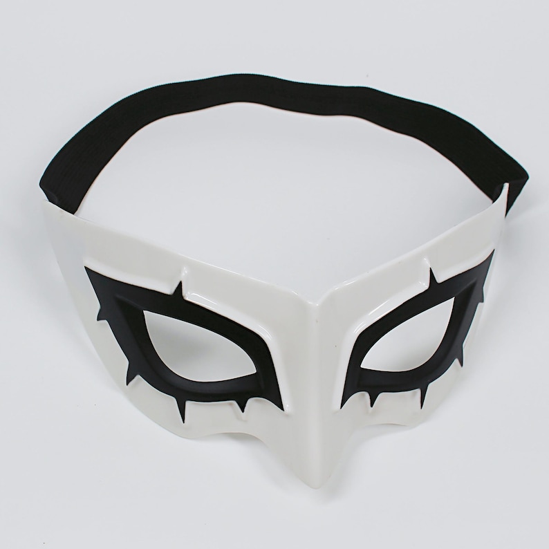 May include: White mask with black eye details and a black elastic band. The mask has a pointed nose and is designed with sharp, angular lines. The eye openings are outlined in black, with stylized points extending outward.
