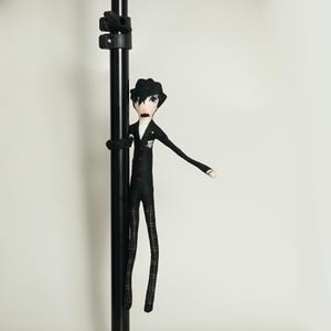 May include: A handmade doll in a black suit with red buttons and plaid trousers, holding onto a black pole. The doll has black hair and a white shirt. The doll's arms are outstretched.