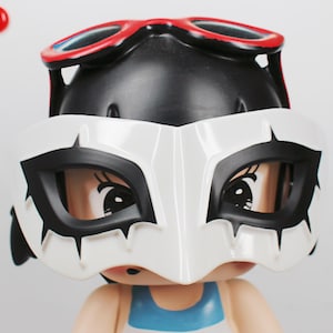 May include: A figurine with a black head, wearing red-rimmed goggles and a white mask with black accents. The figurine has a blue and white top, and the mask covers the eyes, creating a mysterious look.