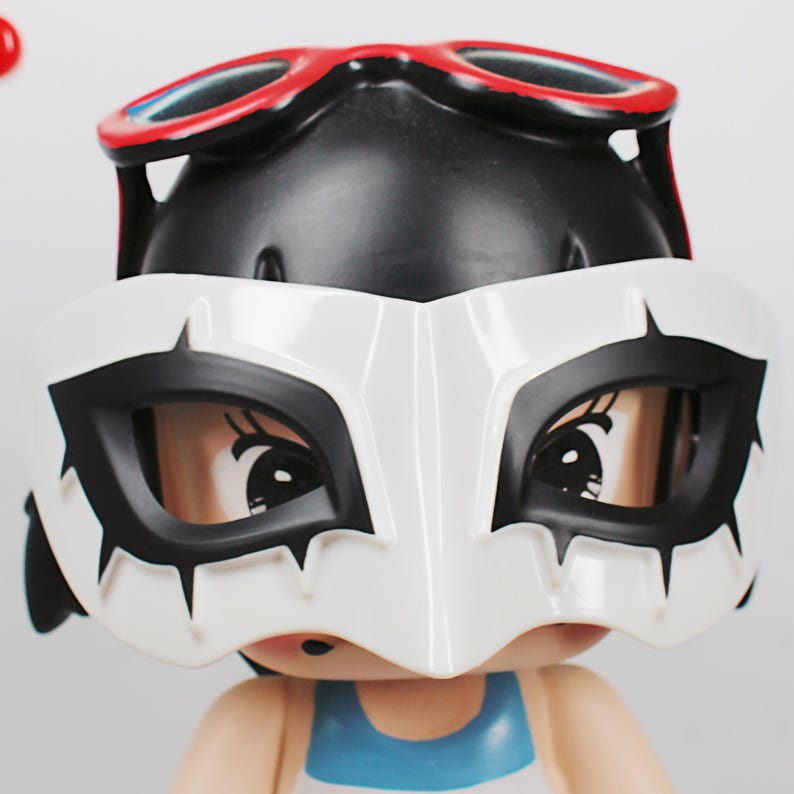 May include: A figurine with a black head, wearing red-rimmed goggles and a white mask with black accents. The figurine has a blue and white top, and the mask covers the eyes, creating a mysterious look.