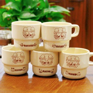 May include: Six beige ceramic mugs stacked together. Each mug has a brown cartoon chef cat design and a name: Rafayel, Sylus, Xavier, Zayne, and Caleb. The mugs are on a wooden surface.