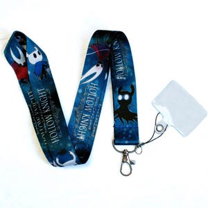 May include: A blue lanyard featuring characters and text from the video game "Hollow Knight." The lanyard has a silver clasp and a clear plastic card holder attached. The design includes white and black character illustrations.