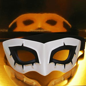 May include: A white and black mask with a black elastic band. The mask has a white base with black eye openings and gold accents. The mask is designed with a pointed shape and is likely for cosplay or costume use.