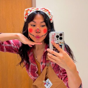 May include: A person wearing a red and black plaid shirt, brown overalls, and a crocheted headband with a scarecrow face painted on their face. They are holding a phone with a floral case and a photo of a person in the case.