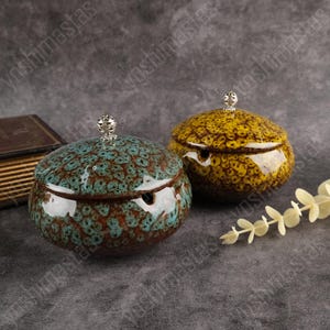 以下が含まれることがあります： 2つの蓋付きセラミックジャー。1つは茶色の斑点模様のターコイズ色、もう1つは同様の模様の黄色です。どちらも上部に銀色のつまみがあります。ジャーは丸いです。