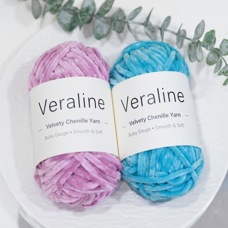 May include: Two skeins of Veraline velvety chenille yarn, one in a soft lavender and the other in a bright turquoise. Each skein is labeled with the brand name and product details: "Velvety Chenille Yarn, Bulky Gauge, Smooth & Soft."