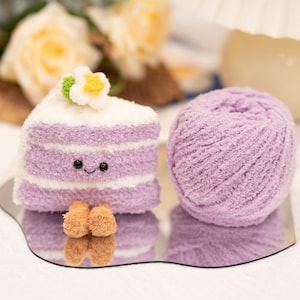 May include: A soft, lilac-coloured cake slice with white frosting and a flower, beside a ball of matching yarn. The cake has a smiling face. The items are on a mirrored surface.
