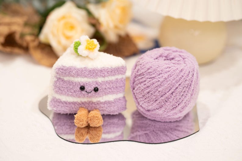 May include: A soft, lilac-coloured cake slice with white frosting and a flower, beside a ball of matching yarn. The cake has a smiling face. The items are on a mirrored surface.
