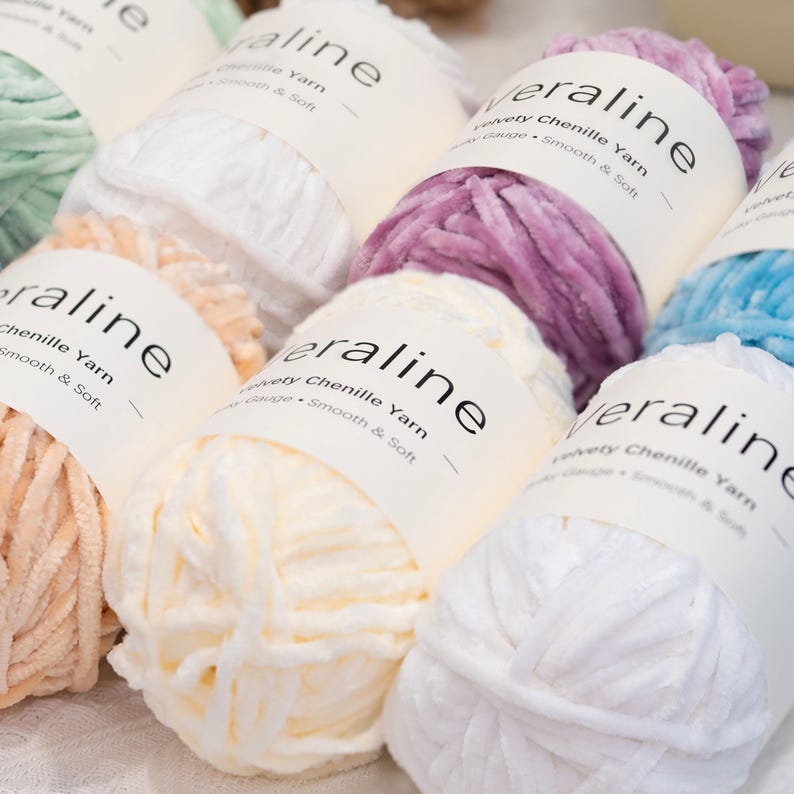 May include: Close-up of several skeins of Veralline chenille yarn in various colors, including white, peach, lavender, and light blue. Each skein has a label with the brand name and yarn details. The yarn appears soft and velvety.