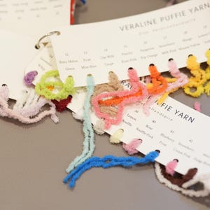 Puede incluir: Primer plano de una carta de colores de hilo con muestras de hilo esponjoso y texturizado en varios colores, incluyendo rosa, amarillo y azul. La carta está etiquetada como "VERALINE PUFFIE YARN" y "J. FIE YARN".