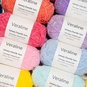 May include: A collection of colorful chenille yarn skeins in various shades, including pink, orange, blue, and yellow. Each skein has a white label with the name "Veraline" and details about the yarn's weight and composition. The yarn is soft and smooth.