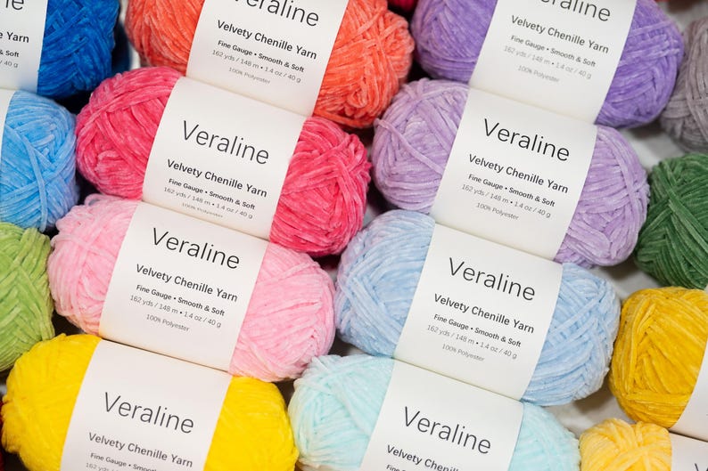 May include: A collection of colorful chenille yarn skeins in various shades, including pink, orange, blue, and yellow. Each skein has a white label with the name "Veraline" and details about the yarn's weight and composition. The yarn is soft and smooth.