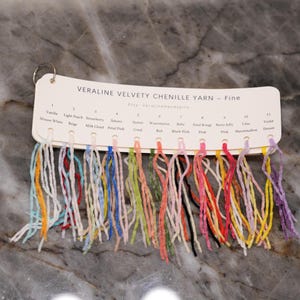 May include: A color chart for "VERALINE VELVETY CHENILLE YARN - Fine" with 11 yarn color samples. Colors include Vanilla, Light Peach, Strawberry, Sakura, Sunset, Watermelon, Baby, Petal Rouge, Berry Jelly, Lilac, and Violet. The chart is on a ring.