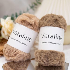 Puede incluir: Dos ovillos de hilo de piel sintética Veraline suave como una pluma. Un ovillo es de color tostado claro y el otro es de color marrón medio. Cada ovillo tiene una etiqueta de papel blanco con la marca Veraline y el texto Feather-Soft Faux Fur Yarn.