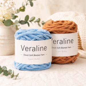 May include: Two skeins of super bulky yarn, one light blue and one terracotta orange, each wrapped in a white paper label reading Veraline Cloud-Soft Blanket Yarn. The yarn has a thick, plush, and textured appearance for crafting projects.