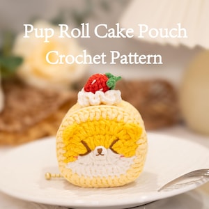 Pode incluir: Uma bolsa de crochê em forma de bolo enrolado, com um design de rosto de cachorro amarelo e branco. É coberto com um morango vermelho e creme branco. O texto "Pup Roll Cake Pouch Crochet Pattern" é exibido acima.