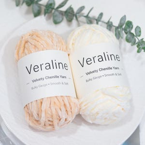 May include: Two skeins of Veraline velvety chenille yarn, one peach and one cream, are displayed on a white plate. Each skein has a white label with the brand name and yarn details: Bulky Gauge, Smooth & Soft.