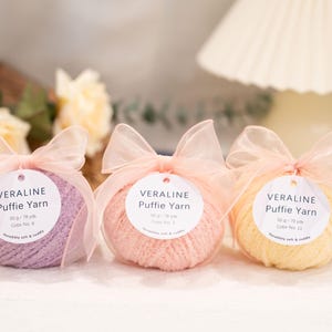 May include: Three balls of Veraline Puffy Yarn in lavender, peach, and yellow, each wrapped in clear plastic and tied with a sheer pink ribbon. Each ball has a white tag with the yarn's name and colour number. Soft and cuddly yarn.