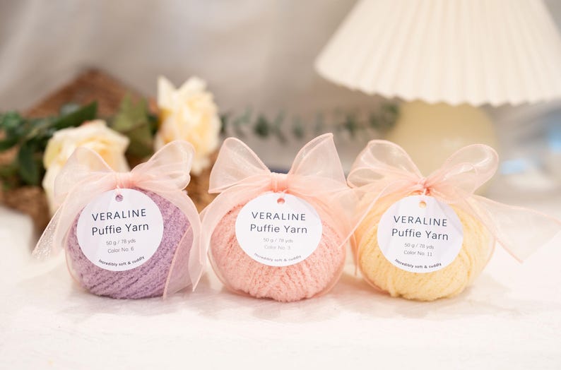 May include: Three balls of Veraline Puffy Yarn in lavender, peach, and yellow, each wrapped in clear plastic and tied with a sheer pink ribbon. Each ball has a white tag with the yarn's name and colour number. Soft and cuddly yarn.