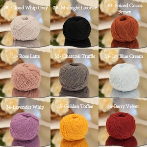 May include: Nine balls of yarn in a range of colours, including grey, black, brown, pink, grey, blue, purple, yellow, and red. Each ball is labelled with a colour name and number. The yarn balls are displayed on a reflective surface.