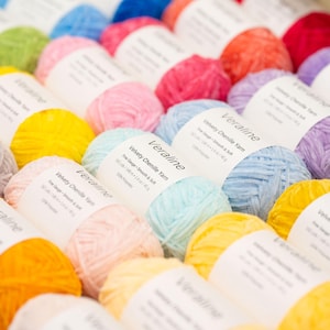 May include: A collection of colorful yarn skeins, including shades of yellow, pink, blue, green, and purple. Each skein is labeled with the text "Veraline Velvety Chenille Yarn". The yarn is arranged in neat rows, creating a visually appealing display for crafting projects.