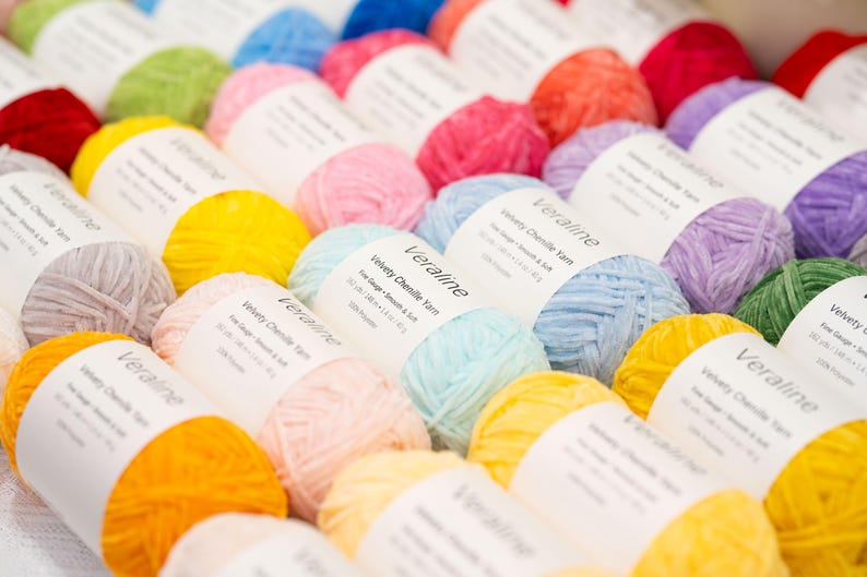 May include: A collection of colorful yarn skeins, including shades of yellow, pink, blue, green, and purple. Each skein is labeled with the text "Veraline Velvety Chenille Yarn". The yarn is arranged in neat rows, creating a visually appealing display for crafting projects.