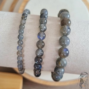 Puede incluir: Tres pulseras de piedra de labradorita gris con cuentas plateadas. Las pulseras están apiladas una encima de la otra sobre una superficie blanca.