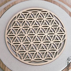 Peut inclure: Une pièce décorative en bois clair, découpée au laser, représentant la Fleur de Vie. Le motif géométrique est composé de cercles qui se chevauchent. Elle est posée sur un fond en tissu blanc, encadrée par un cercle en bois clair.