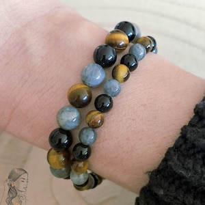 Può includere: Due braccialetti di perline con pietre nere, blu e marroni. I braccialetti sono composti da perline rotonde e vengono indossati al polso. L'immagine include anche il logo "La Pierre Colorée".
