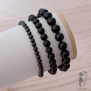 Puede incluir: Tres pulseras de cuentas negras de diferentes tamaños se exhiben sobre un soporte cilíndrico blanco. Las cuentas son redondas y brillantes, reflejando la luz. Las pulseras están dispuestas una al lado de la otra, mostrando los diferentes tamaños de las cuentas.