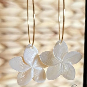 May include: A pair of gold hoop earrings with white, flower-shaped pendants. The earrings are suspended from a black stand against a blurred, textured background. The flower pendants have five petals and a pearlescent sheen.