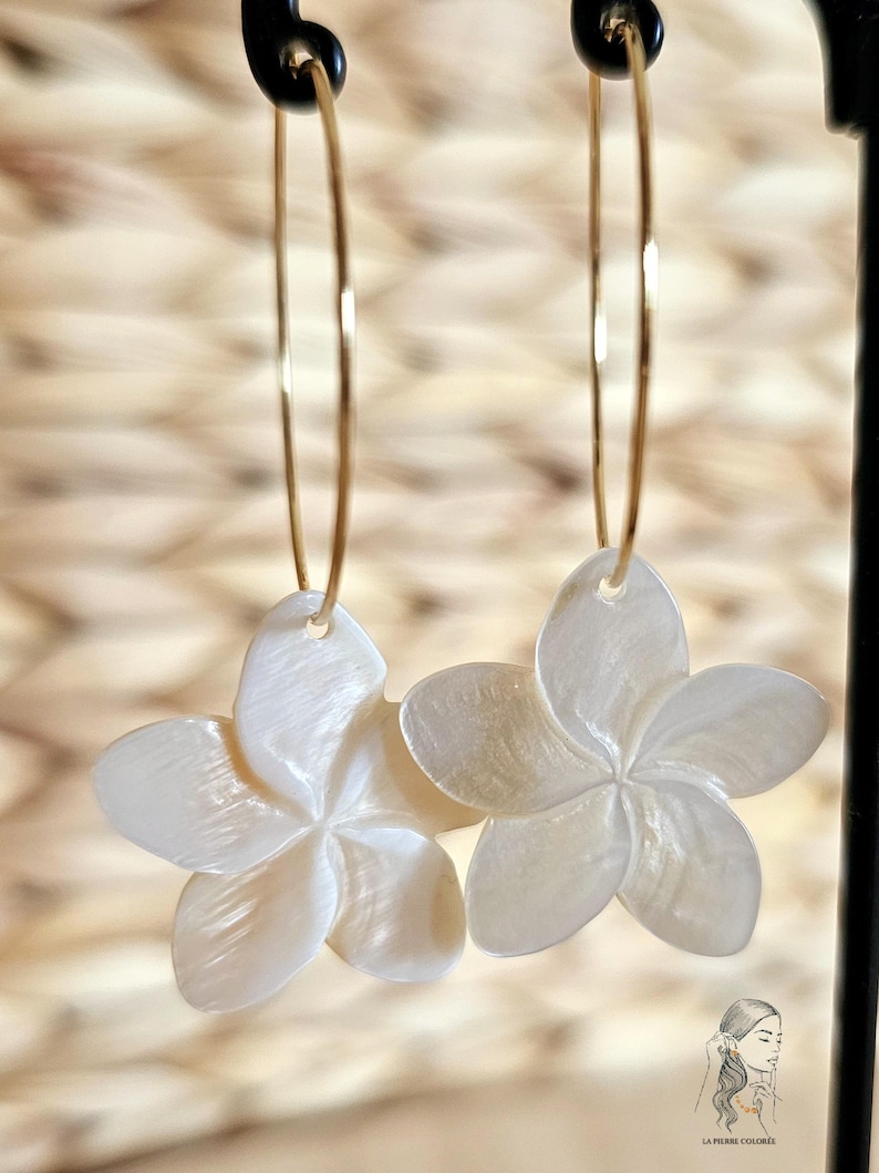 May include: A pair of gold hoop earrings with white, flower-shaped pendants. The earrings are suspended from a black stand against a blurred, textured background. The flower pendants have five petals and a pearlescent sheen.