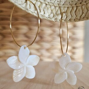 May include: A pair of gold hoop earrings with white, flower-shaped pendants. The earrings are hanging in front of a straw hat and a wooden background. The flower pendants have a pearlescent finish.
