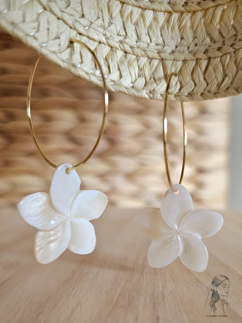 May include: A pair of gold hoop earrings with white, flower-shaped pendants. The earrings are hanging in front of a straw hat and a wooden background. The flower pendants have a pearlescent finish.