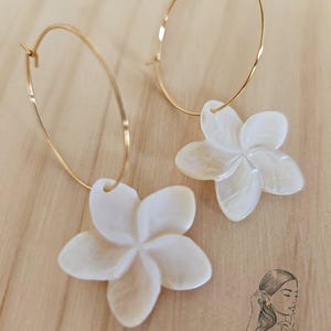 May include: Gold-tone hoop earrings with white, flower-shaped pendants. The earrings feature a delicate design with a polished finish. The flower pendants have a pearlescent sheen, adding a touch of elegance. The earrings are displayed on a light-colored wooden surface.