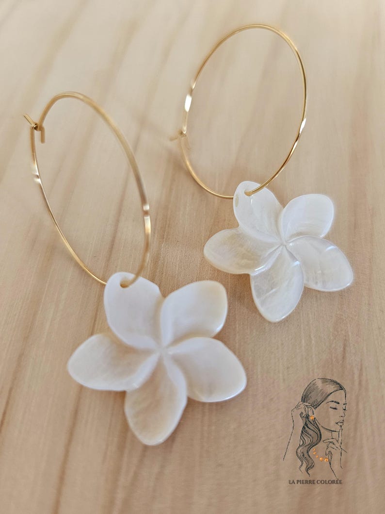 May include: Gold-tone hoop earrings with white, flower-shaped pendants. The earrings feature a delicate design with a polished finish. The flower pendants have a pearlescent sheen, adding a touch of elegance. The earrings are displayed on a light-colored wooden surface.