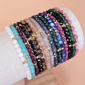 Könnte beinhalten: Eine Sammlung von Armbändern mit Perlen in verschiedenen Farben, darunter Weiß, Türkis, Schwarz und Rosa. Jedes Armband hat kleine, facettierte Perlen und goldfarbene Abstandshalter. Die Armbänder werden auf einer weißen, zylindrischen Form präsentiert.