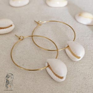 Peut inclure: Boucles d'oreilles créoles dorées avec des coquillages de cauris blancs attachés. Les boucles d'oreilles sont sur un fond blanc.