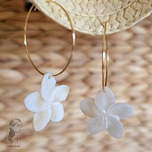 May include: A pair of gold hoop earrings with white flower pendants. The earrings are hanging in front of a woven background. The flower pendants have a detailed petal design.