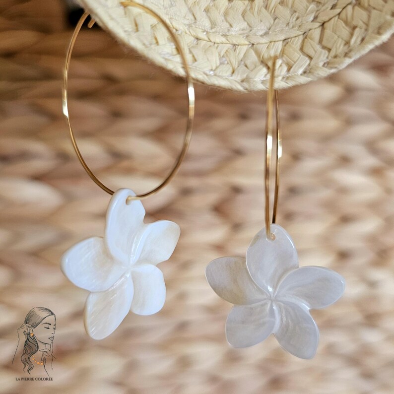 May include: A pair of gold hoop earrings with white flower pendants. The earrings are hanging in front of a woven background. The flower pendants have a detailed petal design.