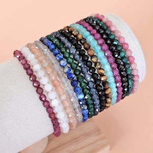 Puede incluir: Una exhibición de pulseras de cuentas en varios colores, incluyendo rojo, blanco, rosa, azul, verde y negro. Las pulseras están hechas de pequeñas cuentas facetadas y se exhiben en un soporte cilíndrico blanco.