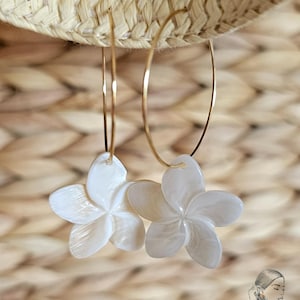 May include: A pair of gold hoop earrings with white, flower-shaped pendants. The earrings are suspended against a woven straw background. The flower pendants have a pearlescent sheen and are detailed with five petals.