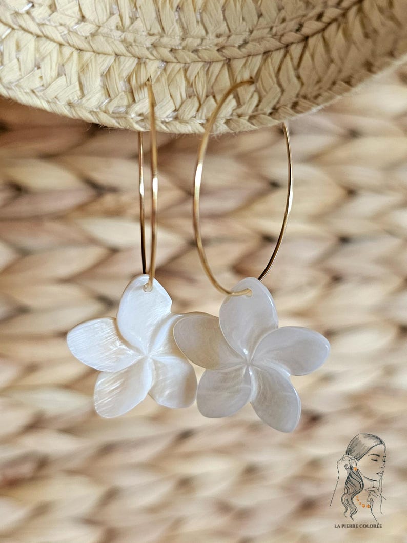 May include: A pair of gold hoop earrings with white, flower-shaped pendants. The earrings are suspended against a woven straw background. The flower pendants have a pearlescent sheen and are detailed with five petals.