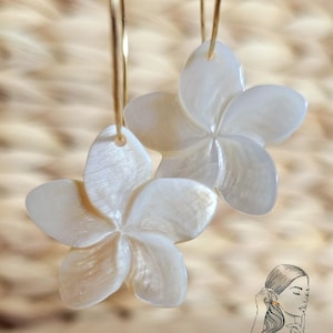 May include: A pair of white flower-shaped earrings with gold-colored hoops. The earrings are made of a pearlescent material and have five petals. The background is a light brown woven texture. The image also includes a small illustration of a woman wearing earrings.