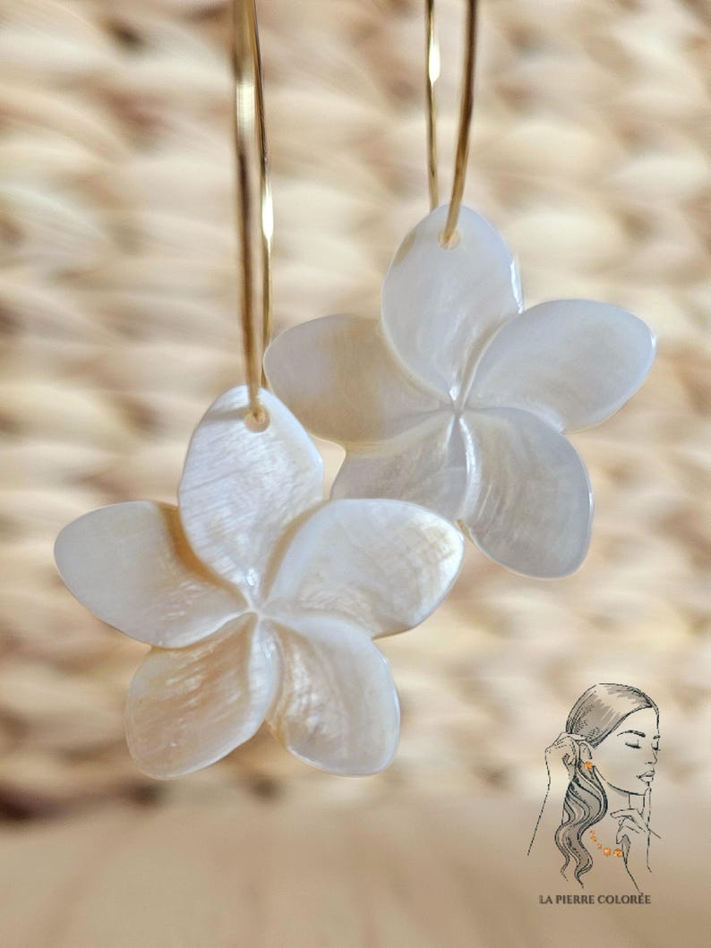 May include: A pair of white flower-shaped earrings with gold-colored hoops. The earrings are made of a pearlescent material and have five petals. The background is a light brown woven texture. The image also includes a small illustration of a woman wearing earrings.
