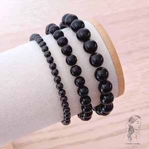 Può includere: Tre braccialetti di perline nere di varie dimensioni sono esposti su un supporto cilindrico bianco. Le perline sono rotonde e hanno una finitura lucida. I braccialetti sono disposti dal più piccolo al più grande, mostrando le diverse dimensioni delle perline.