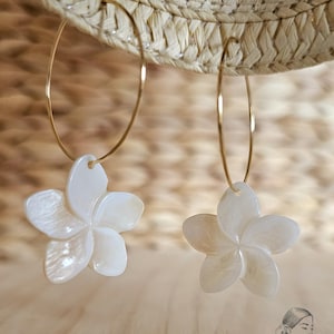 May include: A pair of gold hoop earrings with white, flower-shaped pendants. The earrings are hanging in front of a straw hat. The flower pendants have five petals and a pearlescent finish. The earrings are a jewelry item.