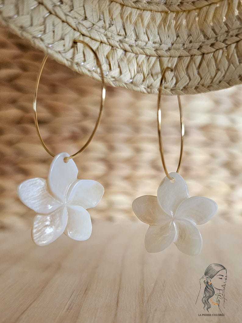 May include: A pair of gold hoop earrings with white, flower-shaped pendants. The earrings are hanging in front of a straw hat. The flower pendants have five petals and a pearlescent finish. The earrings are a jewelry item.