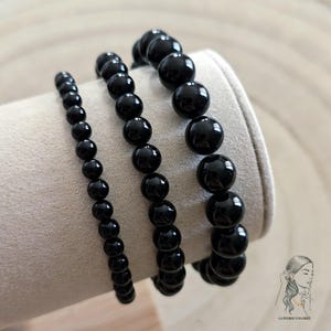 Peut inclure: Trois bracelets de perles noires de tailles différentes sont présentés sur une surface beige. Les perles rondes et brillantes reflètent la lumière. Les bracelets, probablement en onyx, présentent un style de bijou classique. Logo en bas à droite.