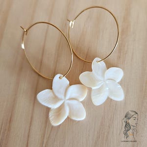 May include: A pair of gold hoop earrings with white, flower-shaped pendants. The earrings are set against a light brown wooden surface. The flower pendants have a pearlescent sheen and are detailed with petal lines.